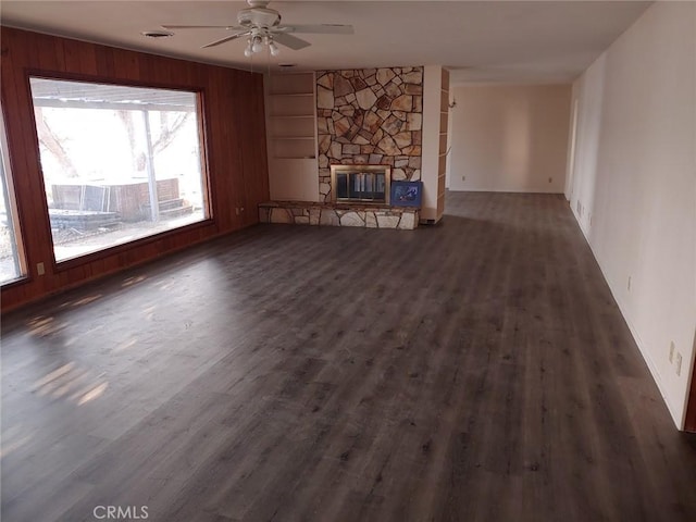 unfurnished living room featuring a stone fireplace, wooden walls, dark hardwood / wood-style flooring, ceiling fan, and built in shelves