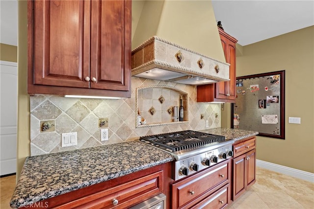 kitchen featuring baseboards, decorative backsplash, custom range hood, dark stone countertops, and stainless steel gas stovetop