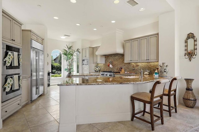 kitchen with light stone countertops, custom exhaust hood, sink, cream cabinets, and built in appliances