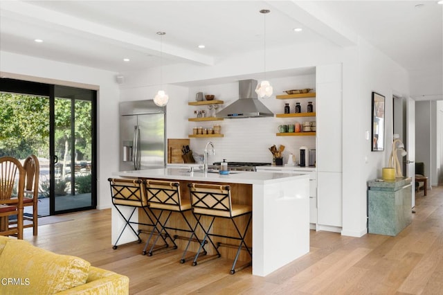 kitchen with beam ceiling, hanging light fixtures, wall chimney range hood, a kitchen breakfast bar, and built in refrigerator