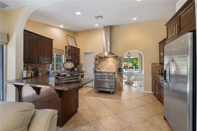 kitchen with light tile patterned flooring, kitchen peninsula, wall chimney range hood, appliances with stainless steel finishes, and a kitchen bar