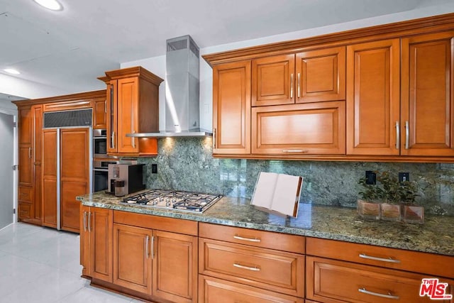 kitchen featuring wall chimney range hood, tasteful backsplash, dark stone countertops, stainless steel gas cooktop, and paneled built in fridge