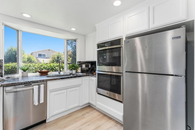 kitchen with appliances with stainless steel finishes, white cabinetry, sink, dark stone countertops, and light hardwood / wood-style flooring