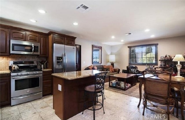 kitchen with a kitchen island, dark brown cabinets, backsplash, light stone countertops, and appliances with stainless steel finishes