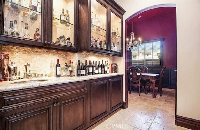 bar with light stone countertops, sink, dark brown cabinets, decorative light fixtures, and a chandelier