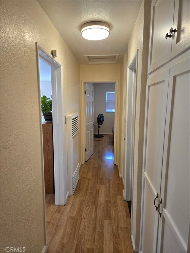 corridor with heating unit, wood finished floors, baseboards, attic access, and a textured wall