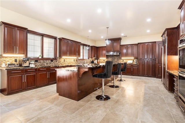 kitchen with pendant lighting, decorative backsplash, a kitchen island, under cabinet range hood, and a kitchen breakfast bar