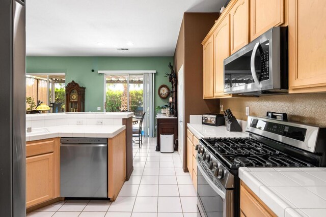 kitchen with appliances with stainless steel finishes, tile countertops, light tile patterned floors, and light brown cabinetry