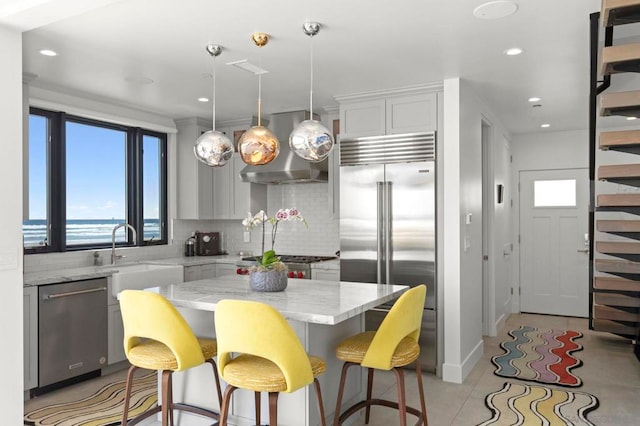 kitchen with wall chimney range hood, appliances with stainless steel finishes, a kitchen island, hanging light fixtures, and decorative backsplash