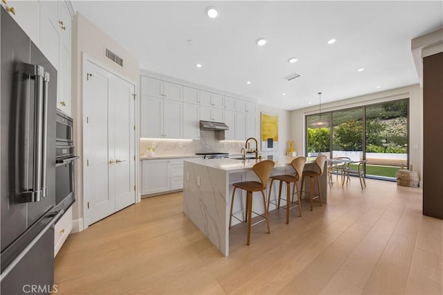 kitchen featuring a center island with sink, light wood-type flooring, decorative light fixtures, white cabinetry, and stainless steel appliances