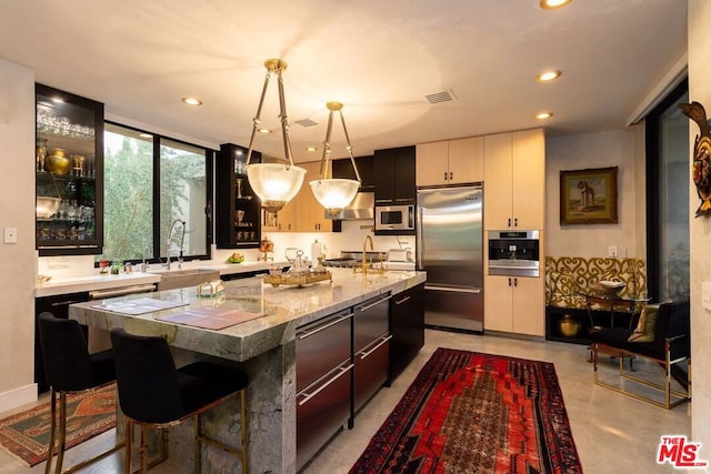 kitchen featuring sink, a kitchen island with sink, stainless steel appliances, light stone countertops, and decorative light fixtures