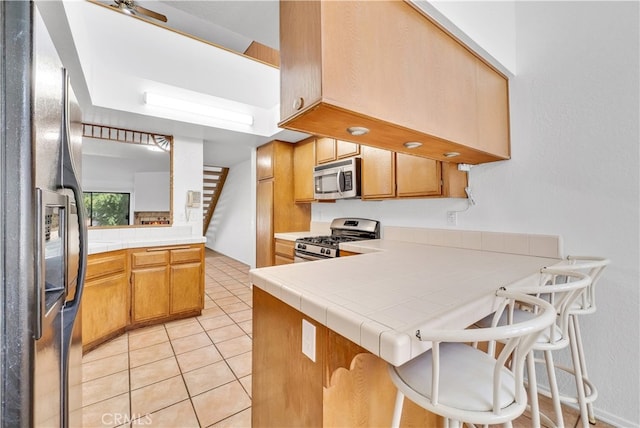 kitchen featuring light tile patterned floors, kitchen peninsula, stainless steel appliances, tile counters, and a kitchen bar