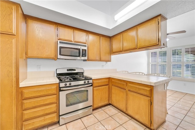 kitchen with appliances with stainless steel finishes, kitchen peninsula, light tile patterned floors, tile countertops, and ceiling fan