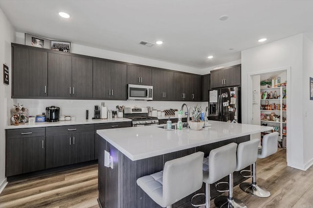 kitchen featuring dark brown cabinetry, an island with sink, wood-type flooring, a kitchen bar, and appliances with stainless steel finishes