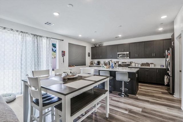 kitchen with appliances with stainless steel finishes, dark brown cabinetry, a center island with sink, hardwood / wood-style flooring, and a breakfast bar area