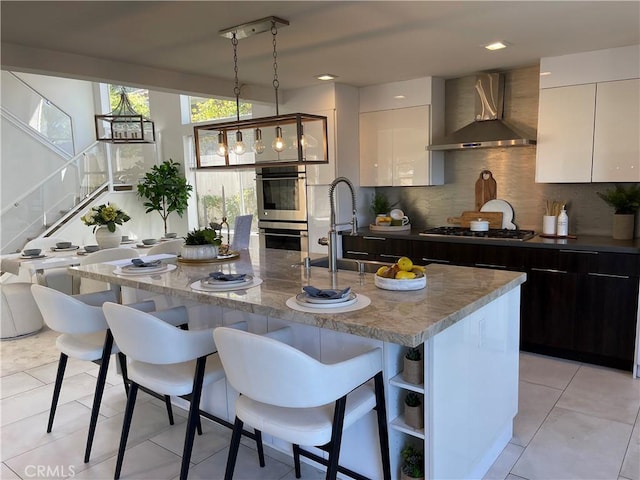 kitchen with dark brown cabinetry, wall chimney exhaust hood, stainless steel appliances, a kitchen island with sink, and white cabinets