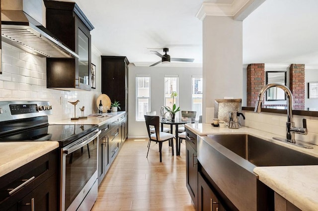 kitchen with stainless steel range with electric cooktop, wall chimney range hood, sink, light wood-type flooring, and ceiling fan