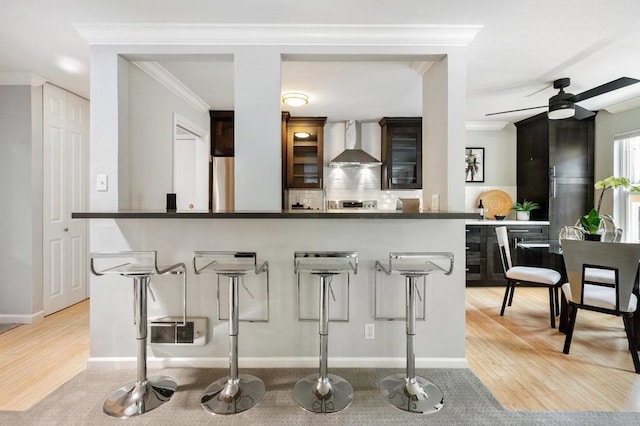 kitchen with ceiling fan, wall chimney exhaust hood, ornamental molding, light hardwood / wood-style floors, and a breakfast bar area