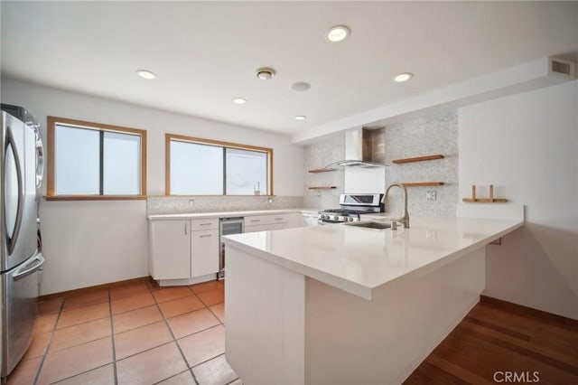 kitchen with sink, white cabinetry, kitchen peninsula, stainless steel appliances, and wall chimney range hood