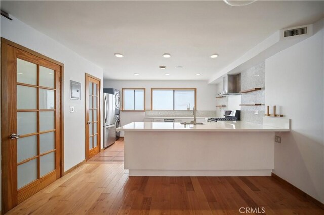 kitchen with stove, kitchen peninsula, stainless steel refrigerator, light wood-type flooring, and wall chimney exhaust hood