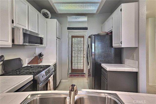 kitchen with baseboards, tile counters, stainless steel appliances, white cabinetry, and a sink