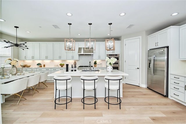 kitchen with a kitchen island with sink, under cabinet range hood, stainless steel appliances, light countertops, and decorative light fixtures