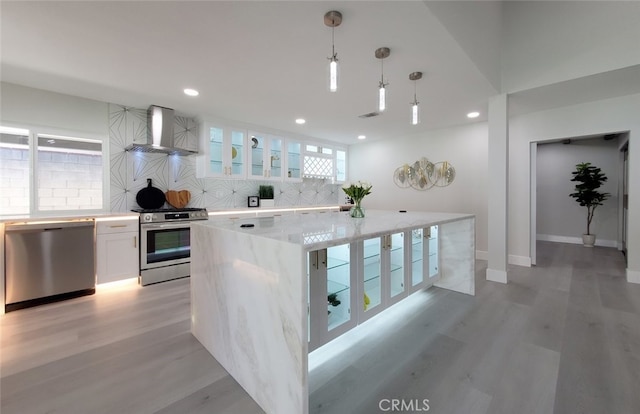 kitchen featuring a center island, white cabinetry, wall chimney range hood, appliances with stainless steel finishes, and decorative light fixtures