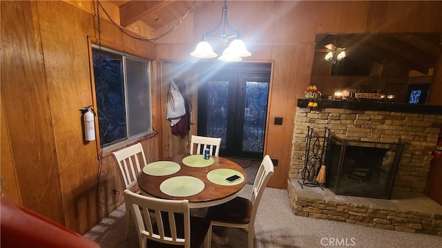 dining area with beam ceiling, carpet flooring, a notable chandelier, wooden walls, and a stone fireplace