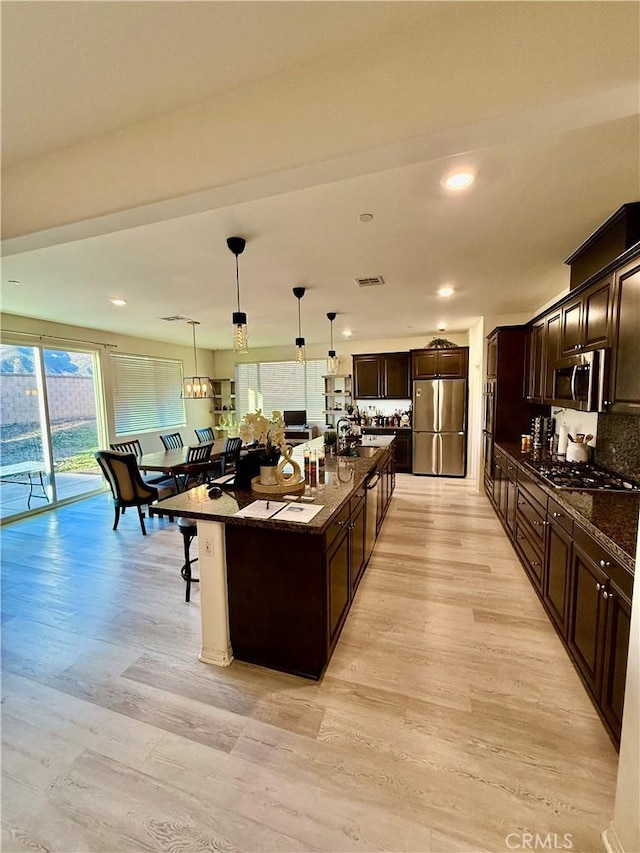 kitchen featuring pendant lighting, light hardwood / wood-style flooring, appliances with stainless steel finishes, a kitchen island with sink, and dark stone countertops