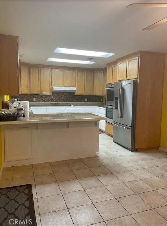 kitchen with appliances with stainless steel finishes, kitchen peninsula, a skylight, ceiling fan, and light tile patterned flooring