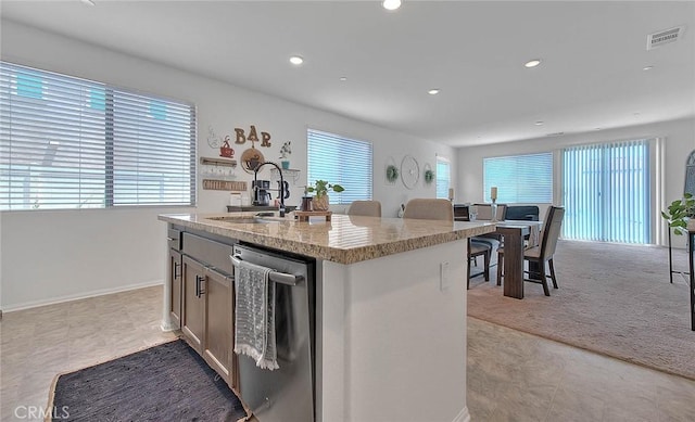 kitchen with light stone counters, light colored carpet, sink, dishwasher, and an island with sink