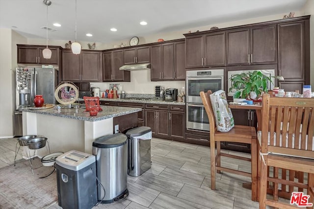 kitchen with appliances with stainless steel finishes, dark stone counters, dark brown cabinets, decorative light fixtures, and an island with sink