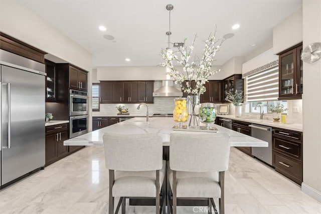 kitchen featuring wall chimney range hood, backsplash, a large island with sink, decorative light fixtures, and appliances with stainless steel finishes