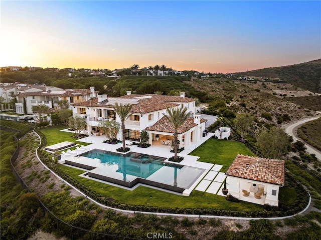 back of house at dusk with a fenced backyard, a lawn, a pool with connected hot tub, and a balcony