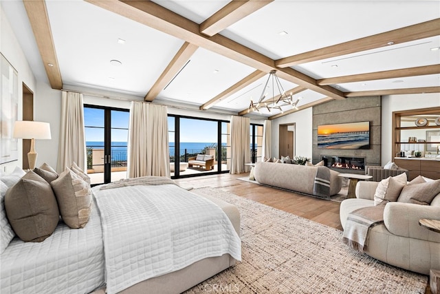 bedroom with lofted ceiling with beams, a water view, light wood-type flooring, a large fireplace, and a notable chandelier