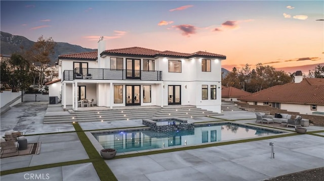 back house at dusk with a balcony, outdoor lounge area, a mountain view, a pool with hot tub, and a patio