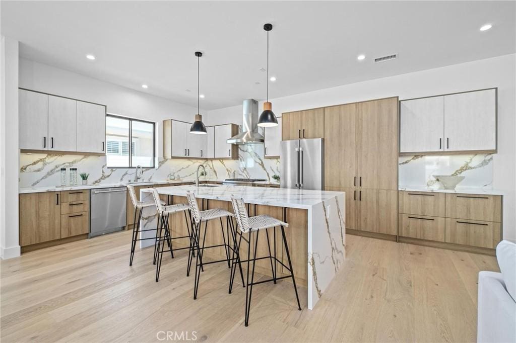 kitchen with a large island with sink, white cabinets, wall chimney range hood, hanging light fixtures, and stainless steel appliances