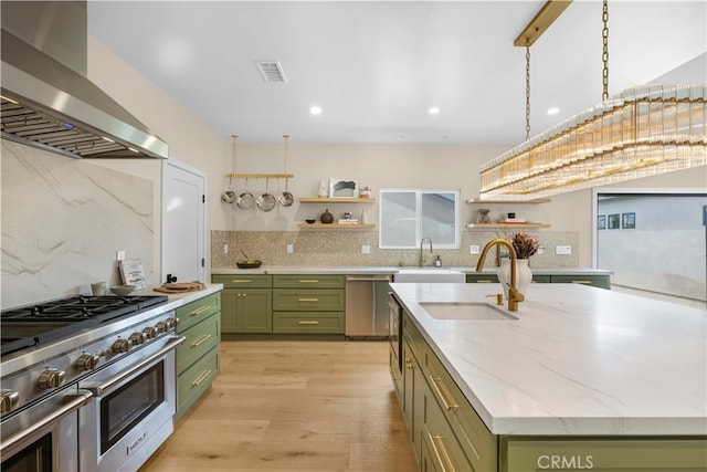 kitchen featuring wall chimney range hood, light hardwood / wood-style flooring, green cabinets, a center island with sink, and appliances with stainless steel finishes