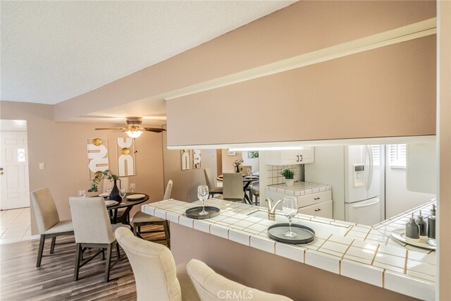 kitchen with tile counters, white fridge with ice dispenser, kitchen peninsula, ceiling fan, and hardwood / wood-style flooring