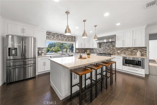 kitchen featuring wall chimney exhaust hood, white cabinets, decorative light fixtures, a kitchen island, and appliances with stainless steel finishes