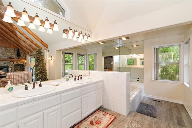 bathroom featuring tiled tub, hardwood / wood-style flooring, vanity, a fireplace, and vaulted ceiling