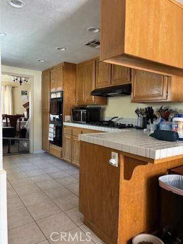 kitchen with light tile patterned floors, double oven, tile counters, kitchen peninsula, and gas cooktop