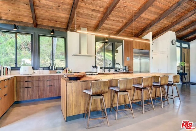 kitchen featuring wall chimney range hood, vaulted ceiling with beams, built in refrigerator, a kitchen island, and wood ceiling