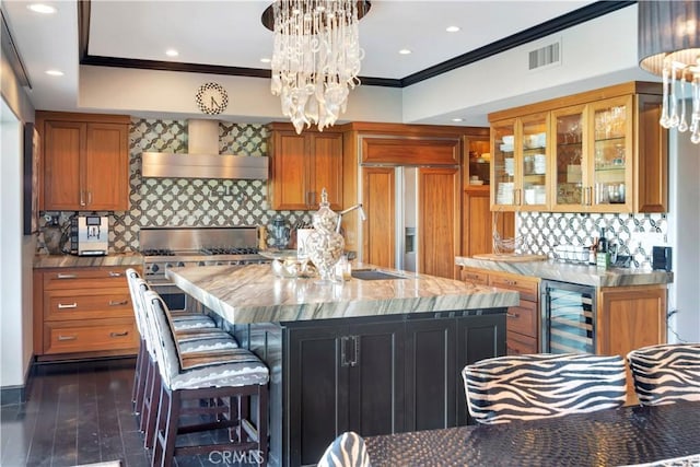 kitchen featuring a kitchen island with sink, wall chimney range hood, tasteful backsplash, decorative light fixtures, and beverage cooler