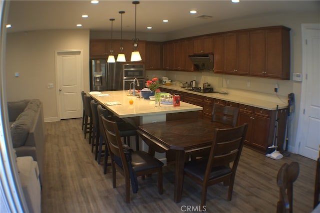 kitchen featuring stainless steel appliances, a kitchen island with sink, dark wood-type flooring, hanging light fixtures, and a breakfast bar area