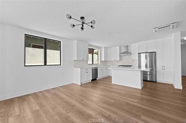 kitchen featuring appliances with stainless steel finishes, white cabinets, a kitchen island, light wood-type flooring, and wall chimney range hood