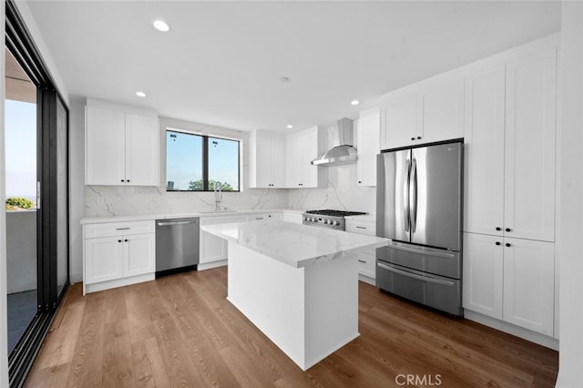 kitchen featuring appliances with stainless steel finishes, white cabinets, wood-type flooring, a center island, and wall chimney range hood