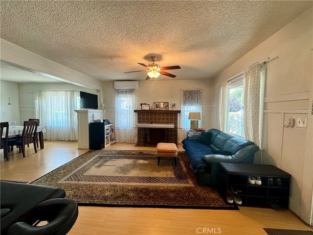 living room featuring ceiling fan, a textured ceiling, a tiled fireplace, light wood-type flooring, and an AC wall unit