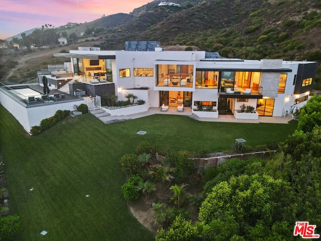 back house at dusk featuring a patio, a mountain view, and a lawn