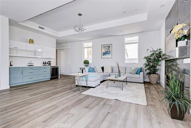 living room with a raised ceiling, wine cooler, a healthy amount of sunlight, and light wood-type flooring